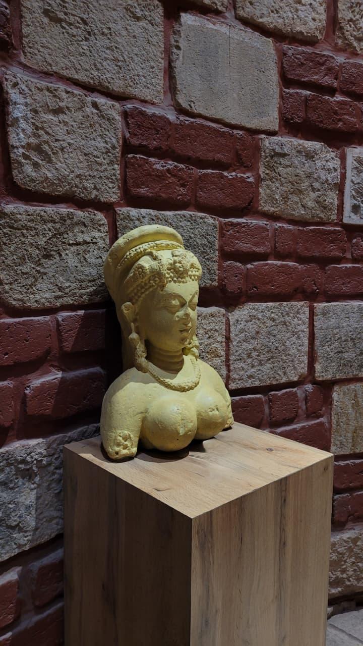 A yellow stone sculpture on a wooden plinth against a brick and stone wall